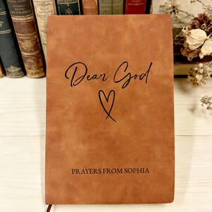 May include: A tan, leather-textured journal featuring the black printed text "Dear God" above a simple heart outline, with the text "PRAYERS FROM SOPHIA" printed in black at the bottom. The cover has a smooth, matte finish and a rectangular shape.