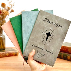 May include: A hand holds a stack of colorful journals. The top journal is gray with the words "Dear God" and "Prayers From Christina" with a cross and dove illustration. Other journals are pink, green, and blue.