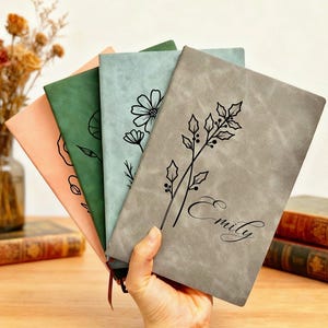May include: A hand holds a stack of four journals in shades of peach, green, blue-green, and gray. Each journal has a floral design and the gray journal is personalized with the name "Emily". The journals are on a wooden surface.
