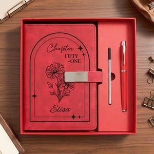May include: A red notebook and pen set in a matching red box. The notebook cover features a black arched frame, a floral illustration, and the text "Chapter FIFTY-ONE" and "Elisa". A silver metal clasp secures the notebook, and a red pen is included.