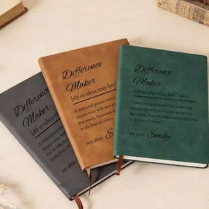 Puede incluir: Tres diarios de cuero en gris, marrón y verde, cada uno con las palabras "Difference Maker" y una definición impresa en la portada. Los diarios tienen marcadores de cinta adjuntos.