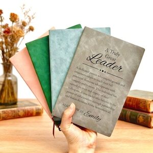 Puede incluir: Un abanico de cuatro cuadernos en gris, azul claro, verde y melocotón. El cuaderno gris presenta el texto A Truly Great Leader, una definición de liderazgo y el nombre Emily impresos en negro. Cada cuaderno tiene un acabado mate y suave.
