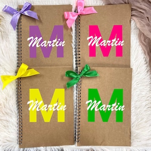 Può includere: Quattro quaderni a spirale con copertine in carta kraft. Ogni quaderno presenta una grande lettera "M" colorata e il nome "Martin" in corsivo bianco. I quaderni sono adornati con fiocchi colorati viola, rosa, gialli e verdi.