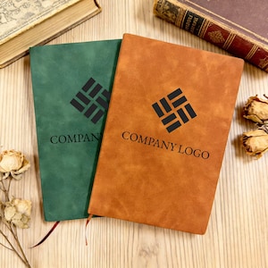 May include: Two notebooks with leather-effect covers, one green and one tan. Each has a black logo and the text "COMPANY LOGO". The notebooks are on a wooden surface, with dried flowers and books in the background.