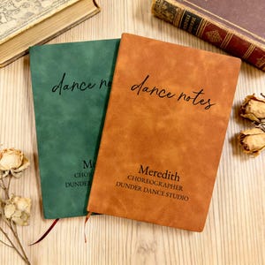 Puede incluir: Dos cuadernos con las palabras "dance notes" en cursiva. Uno es verde, el otro es naranja. El cuaderno naranja tiene el nombre "Meredith" y "CHOREOGRAPHER DUNDER DANCE STUDIO" impresos. Flores secas y libros en el fondo.