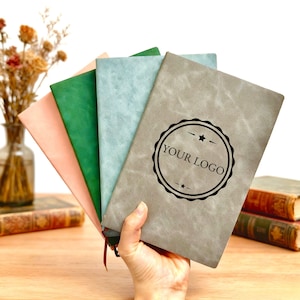 May include: A hand holds a fan of five notebooks in various colors: peach, green, light blue, and gray. The gray notebook has a black circular logo that says "YOUR LOGO". The notebooks have a textured, leather-like appearance.