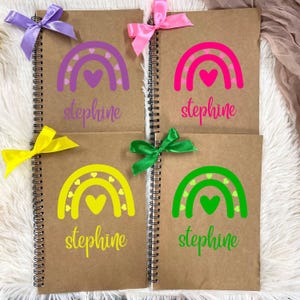 May include: Four spiral-bound notebooks with kraft paper covers. Each notebook features a colorful rainbow design and the name "stephine" in script. The notebooks are adorned with bows in purple, pink, yellow, and green.