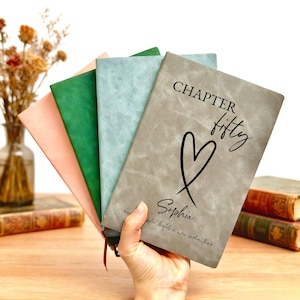 May include: A collection of personalized journals in various colors, including pink, green, and gray. The gray journal features the text "CHAPTER fifty" with a heart and the name "Sophia". Each journal has a ribbon bookmark.