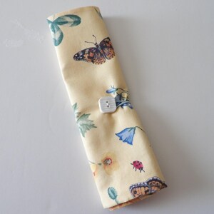 May include: A rolled-up, cream-colored fabric item with a button closure. The fabric is patterned with butterflies, flowers, and leaves in shades of brown, blue, and green. The button is square and white.