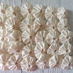 May include: A collection of off-white fabric flowers arranged closely together. Each flower has a circular shape with gathered petals radiating from a central point. The fabric appears to be a natural fiber, possibly cotton or linen.