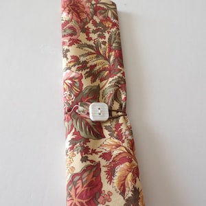 May include: A rolled-up fabric item with a floral pattern in shades of red, green, and beige. It is secured with a white square button and a brown loop. The fabric has a botanical design.