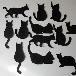 May include: A collection of black cat silhouettes in various poses, including standing, sitting, and stretching. The cats are cut from a dark material and arranged against a white background. The shapes vary in size and posture.