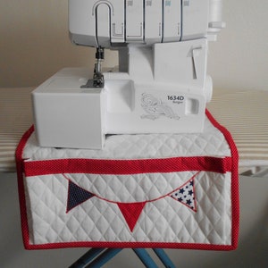 May include: White quilted sewing machine table cover with red and white polka dot trim. The cover has a red, white, and blue bunting design with stars and polka dots. The cover is hanging on a sewing machine table. The sewing machine is a white Singer 1634D Serger.