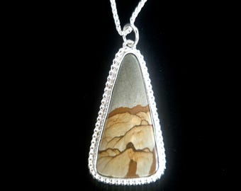 May include: A silver pendant necklace featuring a triangular stone with a landscape design in shades of gray, tan, and brown. The stone is framed by a beaded silver border and hangs from a delicate silver chain. The pendant is set against a black background.