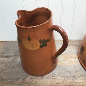 May include: A brown ceramic pitcher with a curved handle and a pouring spout, featuring a painted yellow pumpkin and dark green leaf design around the upper body. A matching brown ceramic bowl with a similar pumpkin motif sits to the right.