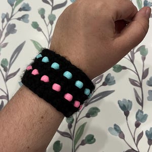 May include: A black crocheted bracelet with pink and blue beads. The bracelet is worn on a person's wrist.