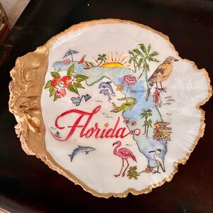 May include: A white seashell dish with a gold rim, painted with a map of Florida, featuring palm trees, a flamingo, a bird, and the word "Florida" in red lettering.