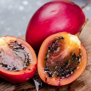 Könnte beinhalten: Nahaufnahme einer Tamarillo-Frucht, auch Baumtomate genannt, aufgeschnitten, um ihre dunklen Samen und ihr orangefarbenes Fruchtfleisch zu zeigen. Die Frucht hat eine leuchtend rote Schale und liegt auf einem Holzbrett. Eine ganze Frucht ist ebenfalls sichtbar.