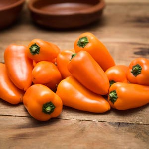 Może przedstawiać: Stos jasno pomarańczowych mini papryczek z zielonymi łodygami na rustykalnej drewnianej powierzchni. Papryki są gładkie i błyszczące. Dwie brązowe miski znajdują się w tle.