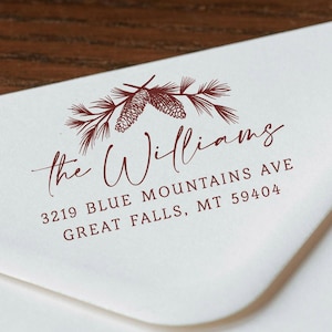 Può includere: Busta bianca con un'illustrazione bordeaux di pigne e aghi. Il testo "the Williams" è scritto in corsivo, con l'indirizzo 3219 Blue Mountains Ave, Great Falls, MT 59404.
