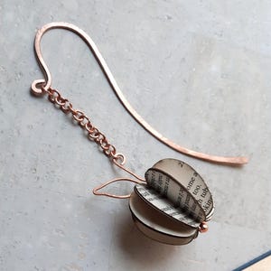 Può includere: Un segnalibro in metallo color rame con un gancio curvo e un ciondolo a forma di mela tridimensionale. La mela è composta da pagine di carta curve e sovrapposte con testo stampato in nero, collegata al gancio tramite una catenella metallica.