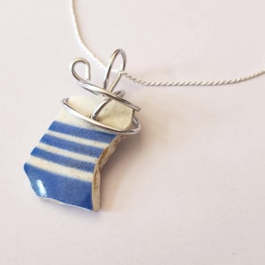 May include: A handmade necklace featuring a piece of blue and white striped ceramic, wrapped in silver wire, and suspended from a silver chain. The ceramic fragment has a nautical aesthetic, reminiscent of coastal decor.