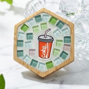 Pode incluir: Um porta-copos hexagonal de bambu com um mosaico de pequenos azulejos de vidro quadrados, verde-claros e transl&uacute;cidos, incrustados em argamassa branca. No centro, uma ilustra&ccedil;&atilde;o gr&aacute;fica de um copo vermelho com palhinha e o texto coke.