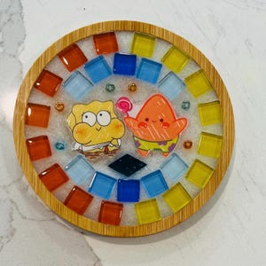 Pode incluir: Um porta-copos circular com moldura de bambu e centro de resina. O design inclui uma esponja amarela e uma estrela-do-mar laranja segurando um pirulito, rodeados por uma borda de azulejos de vidro quadrados e coloridos.