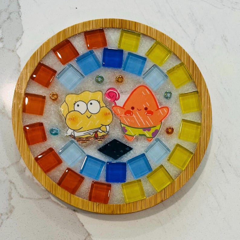 Pode incluir: Um porta-copos circular com moldura de bambu e centro de resina. O design inclui uma esponja amarela e uma estrela-do-mar laranja segurando um pirulito, rodeados por uma borda de azulejos de vidro quadrados e coloridos.