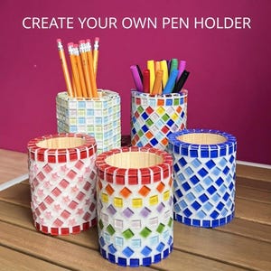 Pode incluir: Cinco porta-canetas cilíndricos decorados com azulejos de mosaico coloridos. O texto CREATE YOUR OWN PEN HOLDER é visível no topo. Os suportes apresentam vários padrões, incluindo quadrados vermelhos, azuis e arco-íris, cheios de lápis.