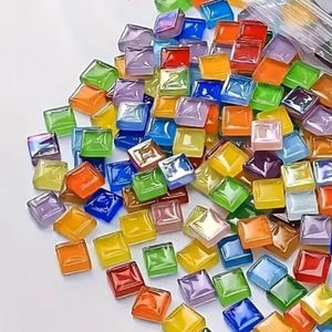 May include: A collection of small, square glass tiles in various colors, including orange, green, blue, yellow, and purple. The tiles are scattered on a white surface, suggesting they are for a mosaic or craft project.