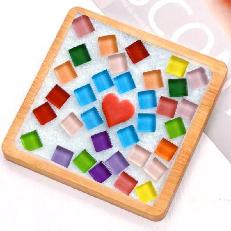 Pode incluir: Um porta-copos quadrado de madeira com uma base branca texturizada incrustada com in&uacute;meros pequenos azulejos de vidro quadrados e coloridos em tons de vermelho, laranja, amarelo, verde, azul e roxo, com um cora&ccedil;&atilde;o de vidro vermelho ao centro.