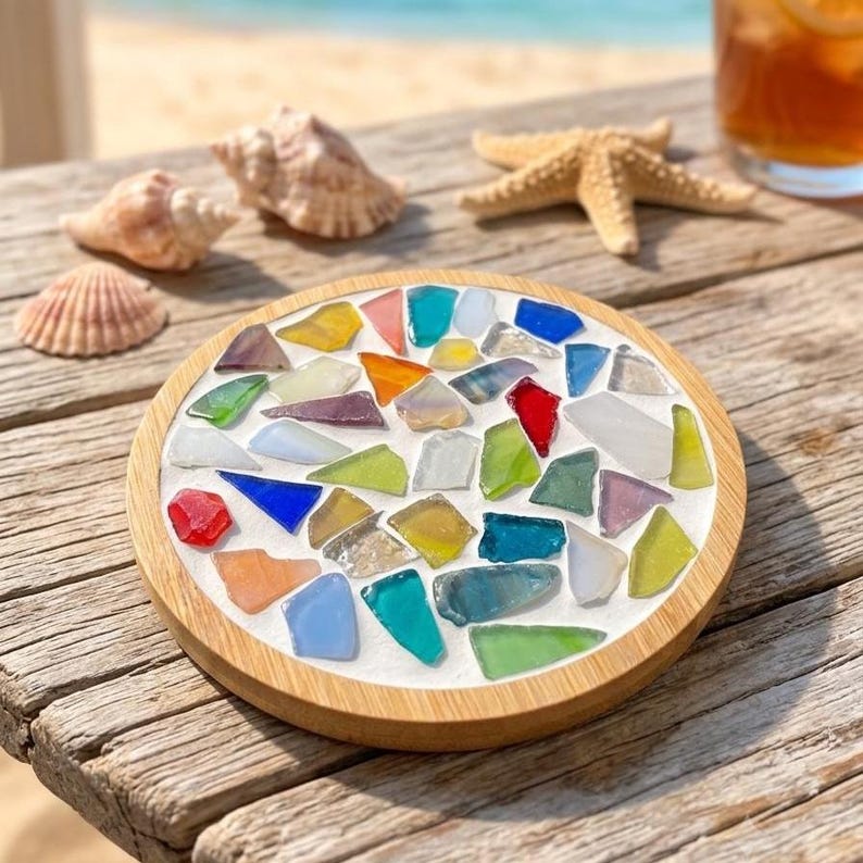 Pode incluir: Um porta-copos circular com uma moldura de madeira clara e uma base branca incrustada com um mosaico de pe&ccedil;as de vidro do mar multicoloridas em tons de azul, verde, amarelo, vermelho e transparente, dispostas num padr&atilde;o disperso na superf&iacute;cie.