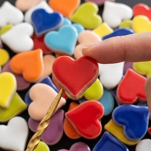 May include: A selection of heart-shaped ceramic tiles in a range of colours, such as red, orange, blue, and white. A red heart tile is held by a finger, highlighting the glossy finish. The tiles are scattered on a dark surface, creating a vibrant display.