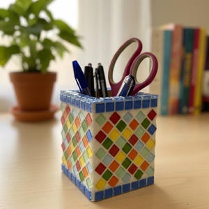 Peut inclure: Un pot à crayons carré en mosaïque avec un motif de losanges colorés. Il contient des stylos et des ciseaux. La mosaïque présente des carreaux bleus, rouges, oranges, jaunes et verts. Le pot est posé sur une surface en bois clair.