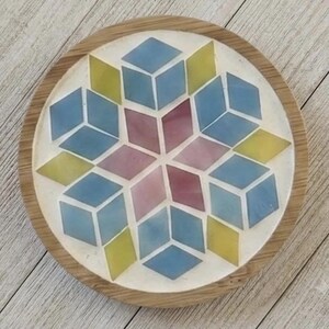 Pode incluir: Um porta-copos circular com uma borda de madeira e um mosaico central. O mosaico consiste em pe&ccedil;as em forma de diamante em tons de azul, amarelo e malva, dispostas num padr&atilde;o geom&eacute;trico de estrela sobre um fundo branco.