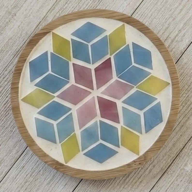 Pode incluir: Um porta-copos circular com uma borda de madeira e um mosaico central. O mosaico consiste em pe&ccedil;as em forma de diamante em tons de azul, amarelo e malva, dispostas num padr&atilde;o geom&eacute;trico de estrela sobre um fundo branco.