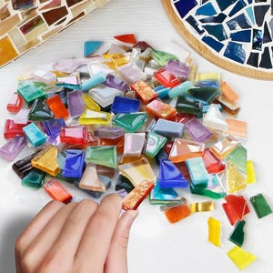 May include: A collection of small, multi-colored glass mosaic tiles in various shapes and sizes, including shades of red, blue, green, yellow, orange, purple, and clear pieces. The tiles are scattered across a surface, with one piece held.