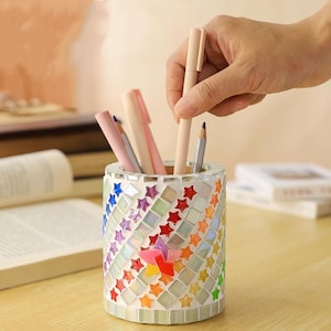 May include: A cylindrical pen holder with a mosaic design. The holder is decorated with colorful star-shaped tiles and square mirrored tiles. A hand is placing a beige pen into the holder, which contains several pens and pencils.