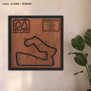 Puede incluir: Una obra de arte mural de madera que representa el diseño del circuito de Road America. La obra tiene un marco oscuro, un fondo de madera marrón claro y líneas de pista negras. El texto "ROAD AMERICA" y "ELKHART LAKE, WISCONSIN" son visibles.