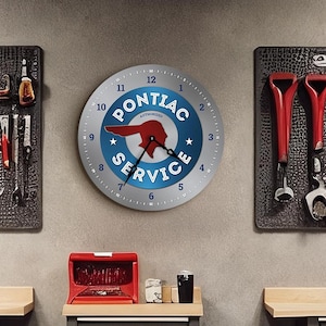 Op de afbeelding: Een werkplaats met een Pontiac Service klok, gereedschapsorganizers en grijze gereedschapskasten met rode accenten. De klok heeft een blauw en rood logo. Gereedschappen zijn netjes gerangschikt op getextureerde wandpanelen, en de kasten hebben houten werkbladen.