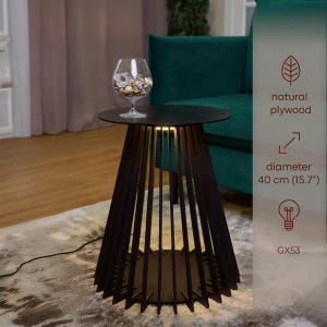 May include: A dark brown side table with a round top and a unique slatted design. The table has a diameter of 15.7 inches (40 cm). A glass bowl filled with decorative items sits on top. The table is illuminated from within.