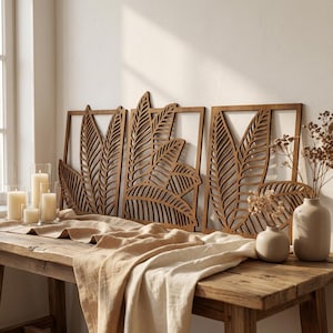 Può includere: Tre pannelli di arte murale in legno con un design a foglie. I pannelli sono incorniciati e hanno una finitura in legno naturale. I pannelli sono esposti sopra un tavolo di legno con candele, vasi e un runner beige.