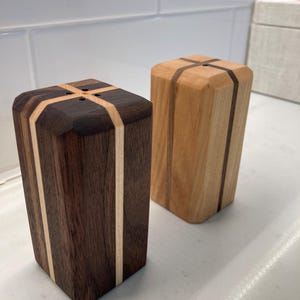 May include: Two wooden salt and pepper shakers. One is dark brown with light wood accents, the other is light brown with dark wood accents. Both have a cross design on top and are rectangular in shape.