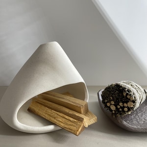 May include: A white speckled ceramic incense holder with a curved, conical shape containing several light brown wooden sticks, positioned next to a dark grey textured dish holding a bundle of dried green herbs wrapped in white twine.