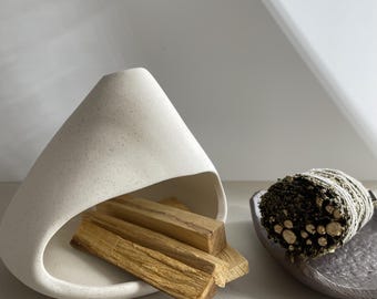 May include: A white speckled ceramic incense holder with a curved, conical shape containing several light brown wooden sticks, positioned next to a dark grey textured dish holding a bundle of dried green herbs wrapped in white twine.