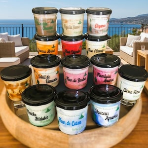 Peut inclure: Une sélection de pots en verre avec des couvercles noirs, chacun portant un nom de parfum différent en français. Les pots sont disposés sur un plateau en bois, avec une vue panoramique sur l'océan en arrière-plan. Les étiquettes incluent "Tiquila Suave", "Elégance", et "Fleur de Coton".