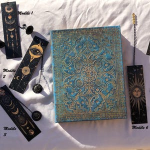 May include: A blue and gold embossed journal with matching bookmarks. The journal has an intricate pattern. The bookmarks are black with celestial designs and dangling charms. The text "Modèle" is visible on the bookmarks.