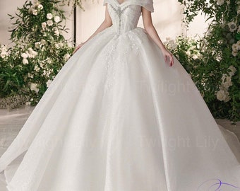 May include: A white off-the-shoulder wedding dress with a full skirt and beaded embellishments. The dress is photographed in front of a floral backdrop.