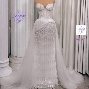 Puede incluir: Un vestido de novia blanco sin tirantes con escote corazón, adornado con perlas y cristales. El corpiño ajustado se transforma en una falda en capas con una larga cola. El vestido se exhibe en un maniquí. Los textos "Twilight Lily" y "Novi 1" son visibles.